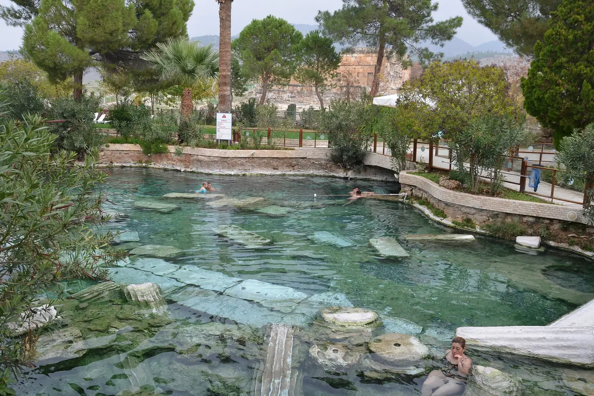 The Antique Pool at Hierapolis, a clear green thermal pool with fallen white marble column pieces lying on its floor, a few swimmers, and trees and ruins behind