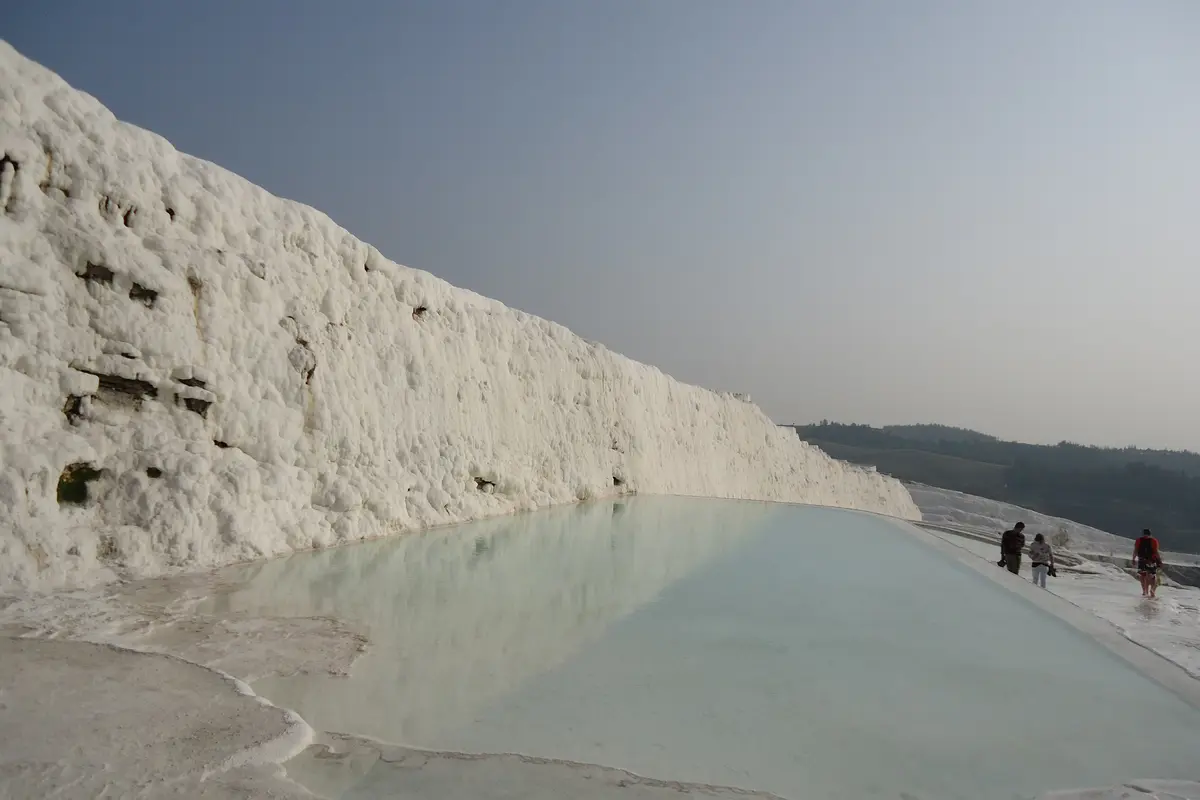 A long pale-blue travertine pool beside a wall of white calcite at Pamukkale, with three visitors walking barefoot along the white path to the right